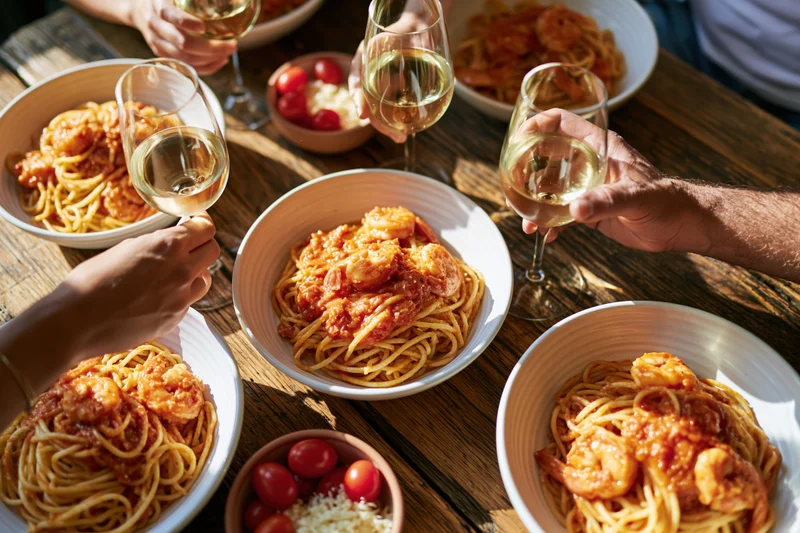 Hands toasting with wine glasses over bowls of shrimp spaghetti on a wooden table