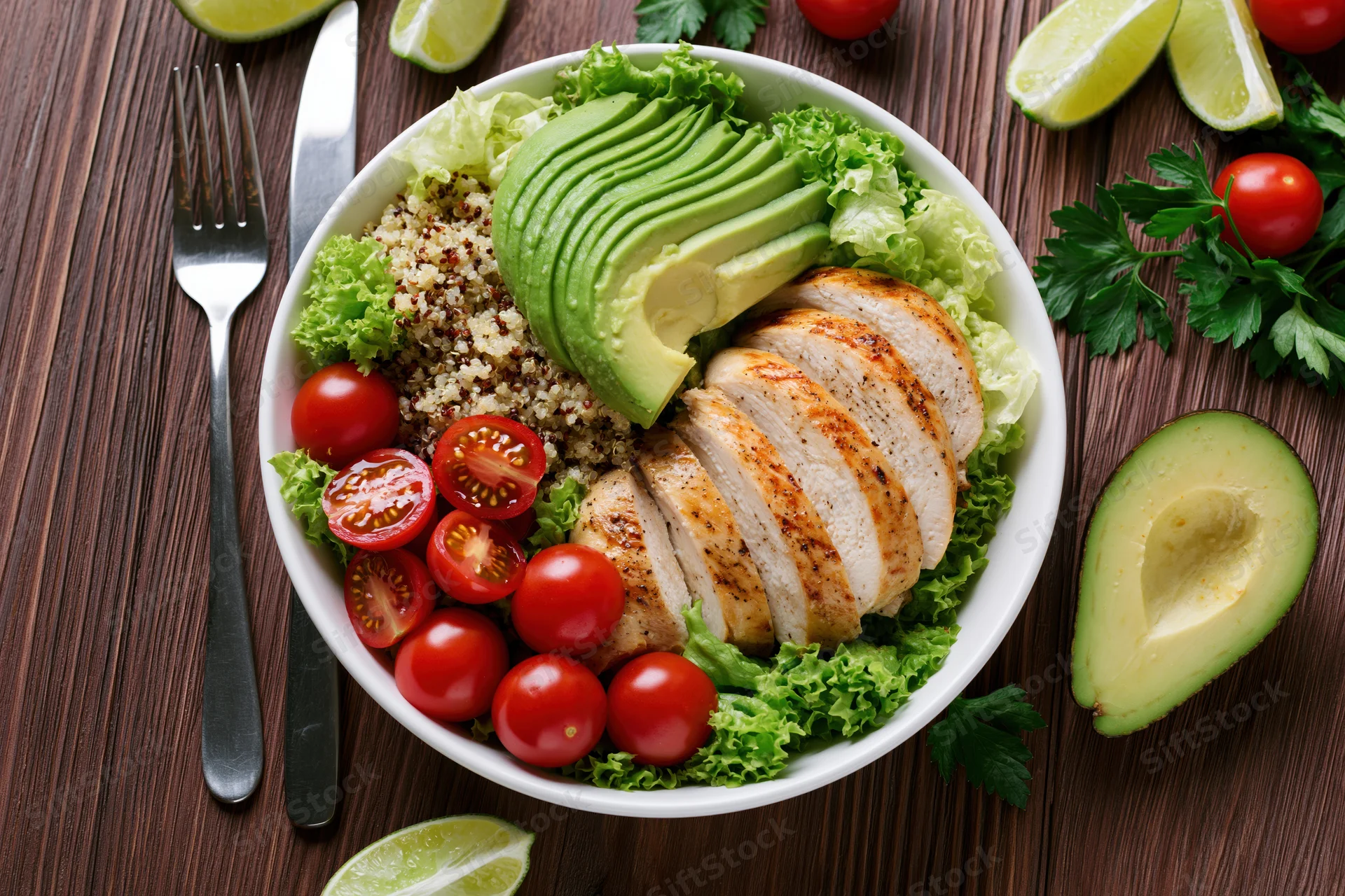 Overhead view of a white bowl filled with sliced chicken, avocado, quinoa, cherry tomatoes and lettuce on a wooden table with cutlery and garnishes around