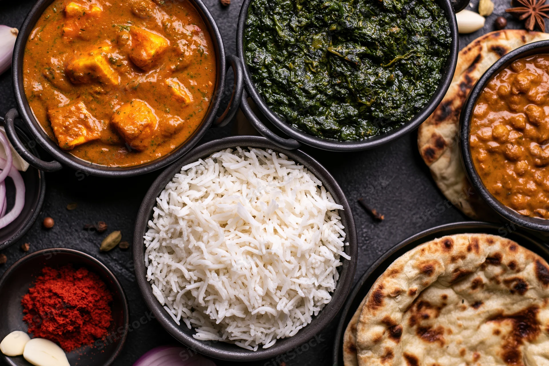 Overhead flat-lay of indian meal with paneer curry, spinach, dal, rice, naan, and spices arranged on dark surface