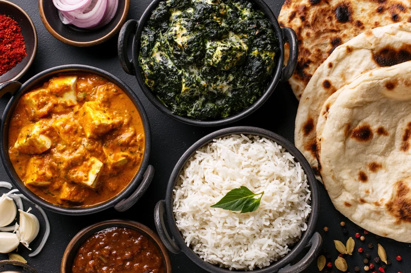 Overhead flat-lay of indian meal with paneer butter chicken, palak paneer, rice, naan, and spices on dark surface