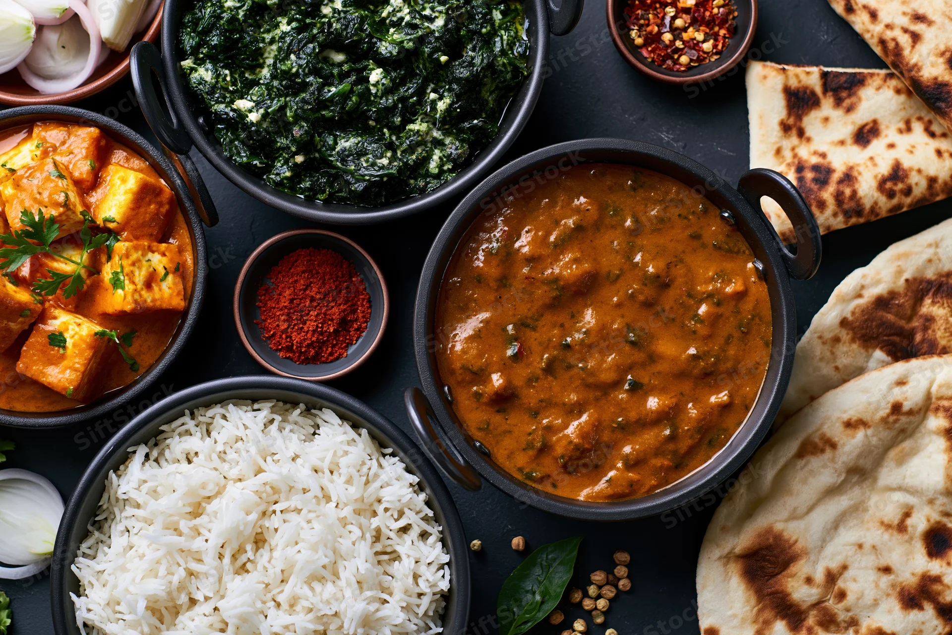 Overhead flat-lay of indian meal with paneer curry, spinach, rice, and naan bread on dark surface