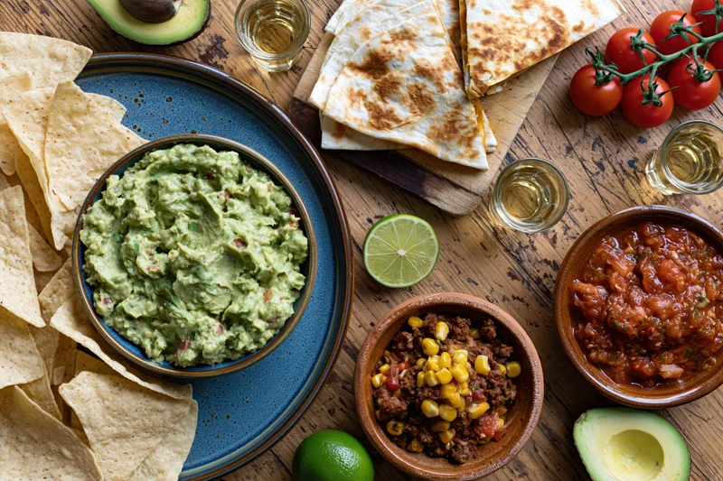 Overhead flat-lay of mexican food spread on a worn wooden table with guacamole, tortilla chips, salsa, lime, tomatoes, avocado, corn beef mix, and tequila glasses