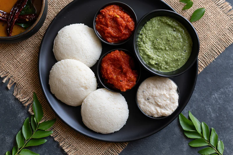 Overhead flat-lay of south indian idli breakfast with chutney and sambar on a black plate