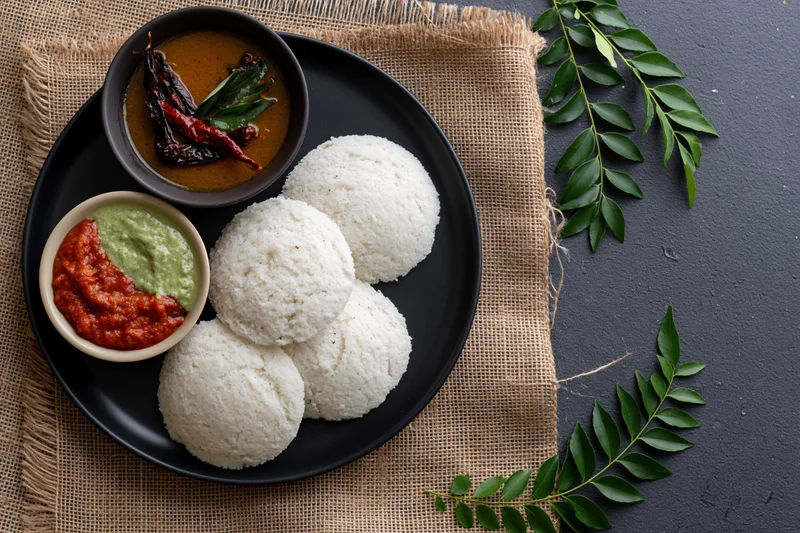 Five white idlis served on a black plate with chutney and curry on a burlap cloth
