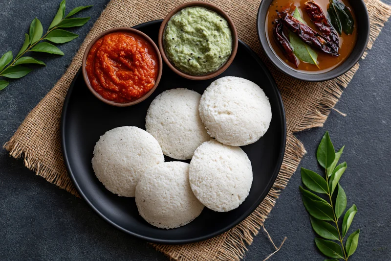 Five white idlis served on a black plate with red and green chutney and sambar, arranged on burlap cloth with curry leaves