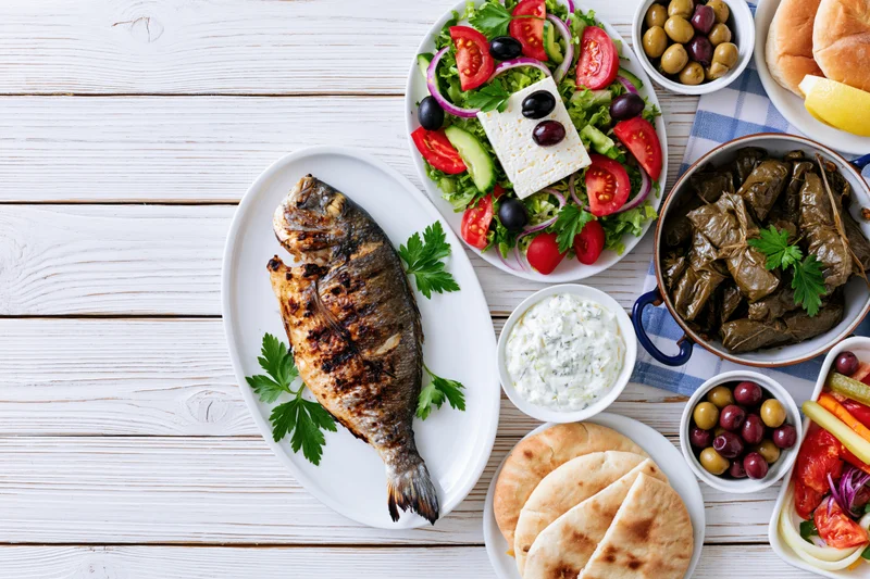 Grilled fish and greek mezze spread arranged on white wooden table with salad, dolmades, pita, and tzatziki