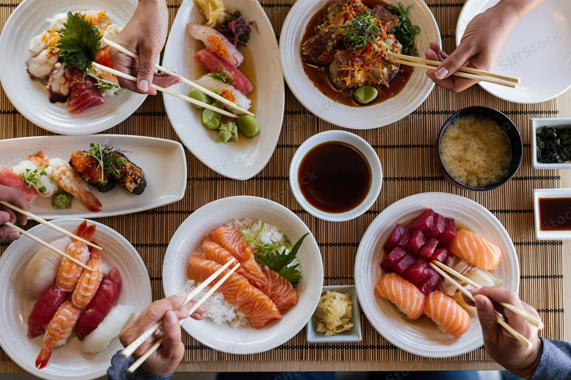 Overhead view of a Japanese restaurant table with multiple plates of sushi and sashimi being eaten with chopsticks by several people sharing a meal on a bamboo mat