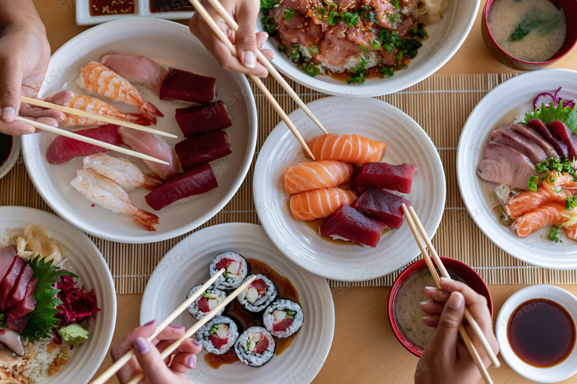 Overhead view of a Japanese restaurant table with multiple plates of sushi and sashimi being eaten with chopsticks by several people