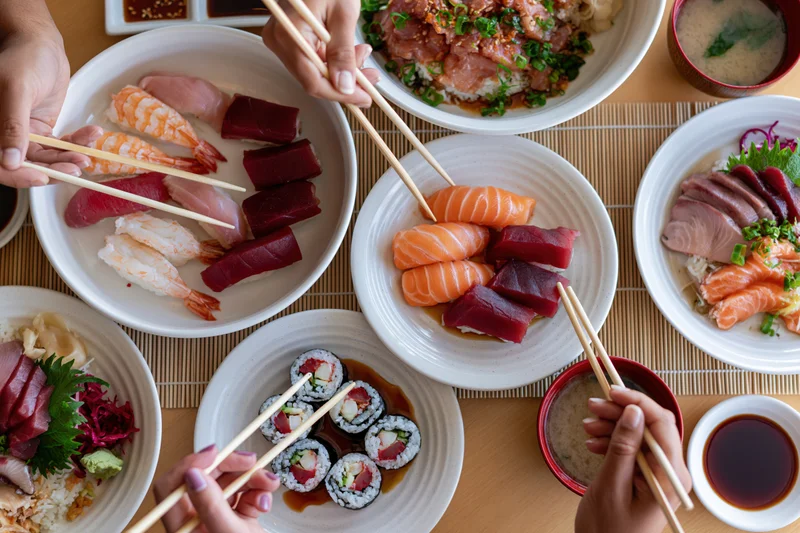 Overhead view of a Japanese restaurant table with multiple plates of sushi and sashimi being eaten with chopsticks by several people