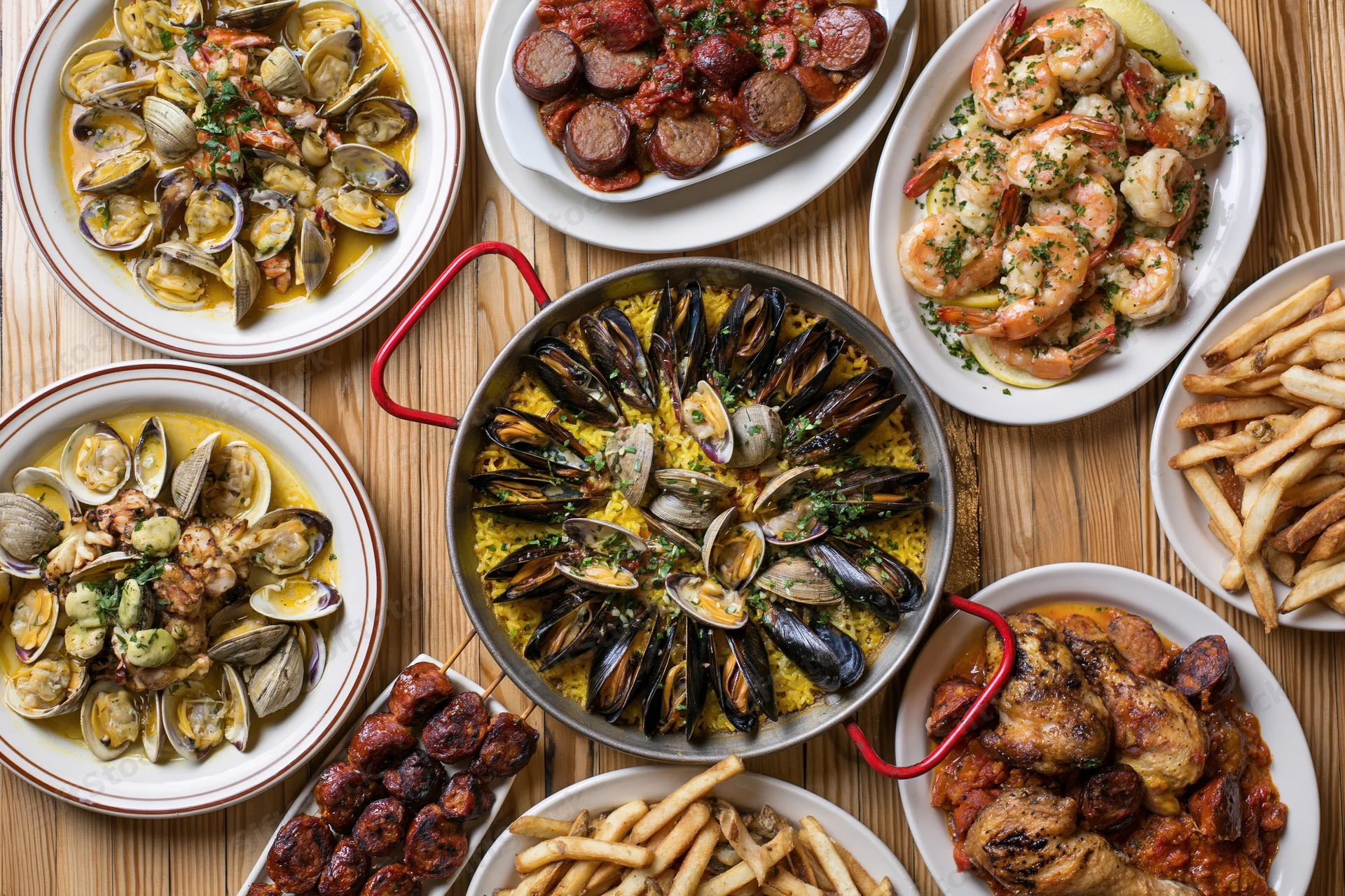 Overhead flat-lay of a Portuguese seafood spread with paella, mussels, shrimp, sausage, fries and clams on a wooden table