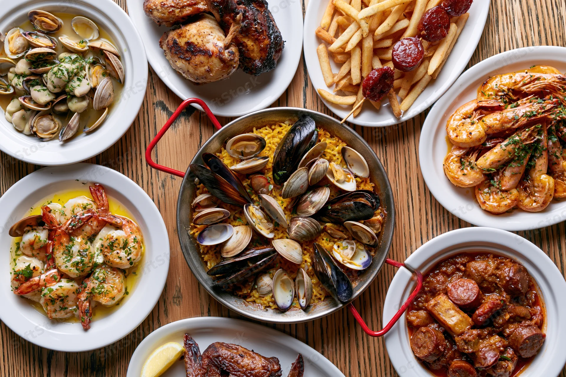 Overhead flat-lay of a portuguese seafood spread on a wooden table with multiple dishes including clams, mussels, shrimp, chicken, fries, and sausage