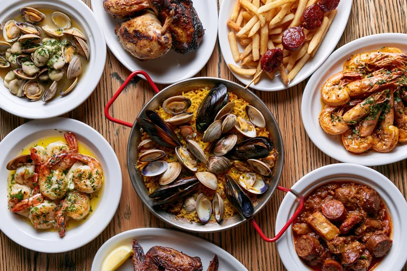 Overhead flat-lay of a portuguese seafood spread on a wooden table with multiple dishes including clams, mussels, shrimp, chicken, fries, and sausage
