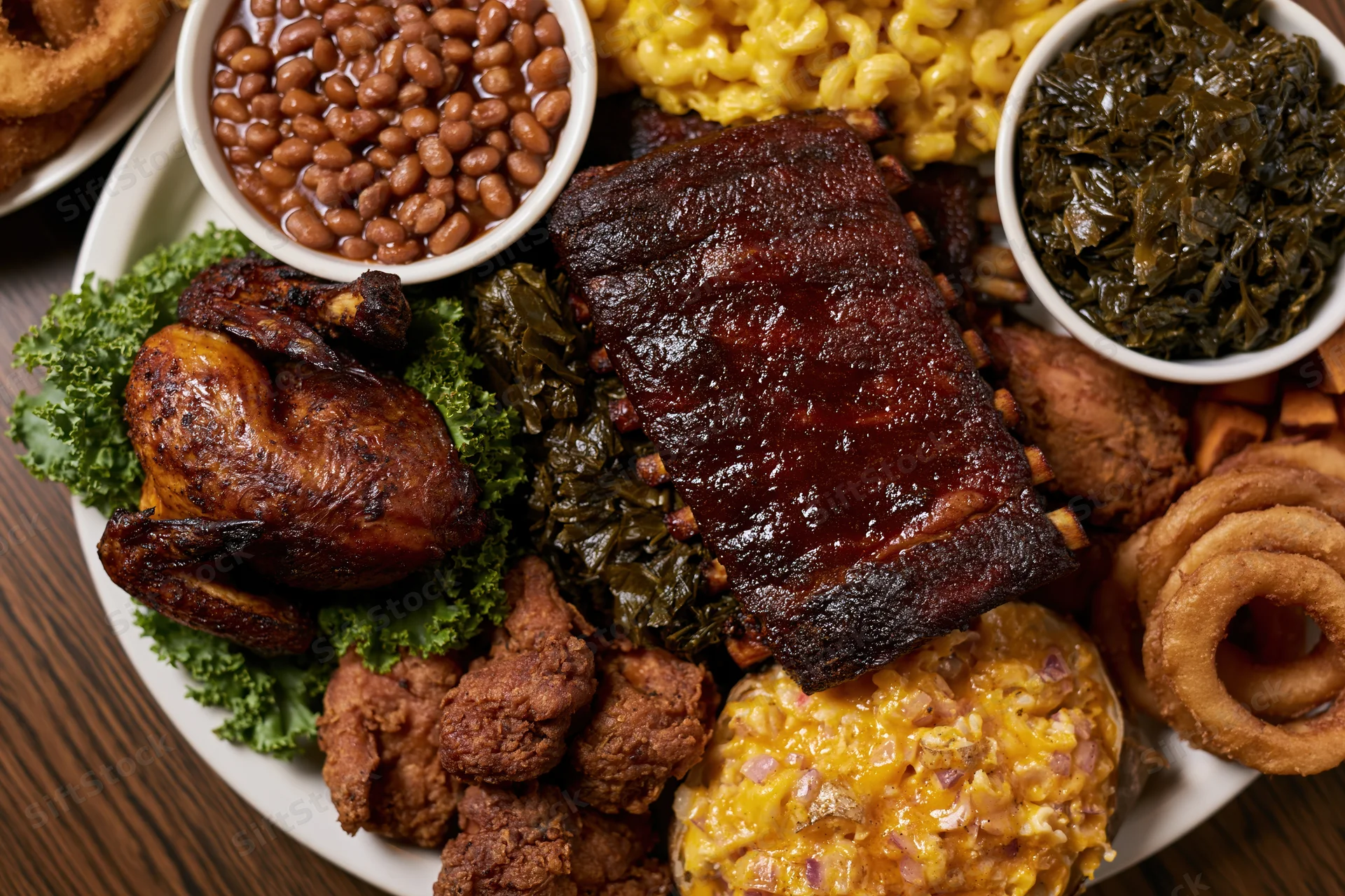 Overhead view of a southern barbecue meal with ribs, chicken, macaroni and cheese, beans, collard greens, onion rings, and fried chicken on a white plate over a dark wooden table