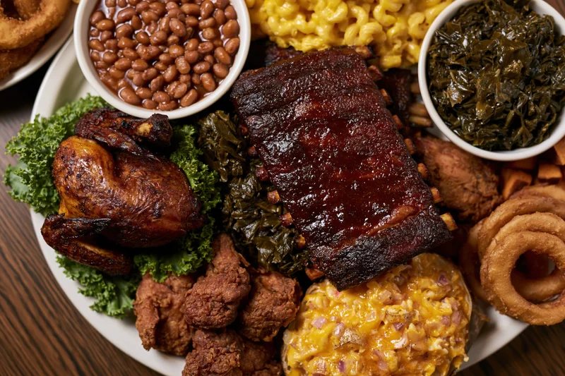Overhead view of a southern barbecue meal with ribs, chicken, macaroni and cheese, beans, collard greens, onion rings, and fried chicken on a white plate over a dark wooden table