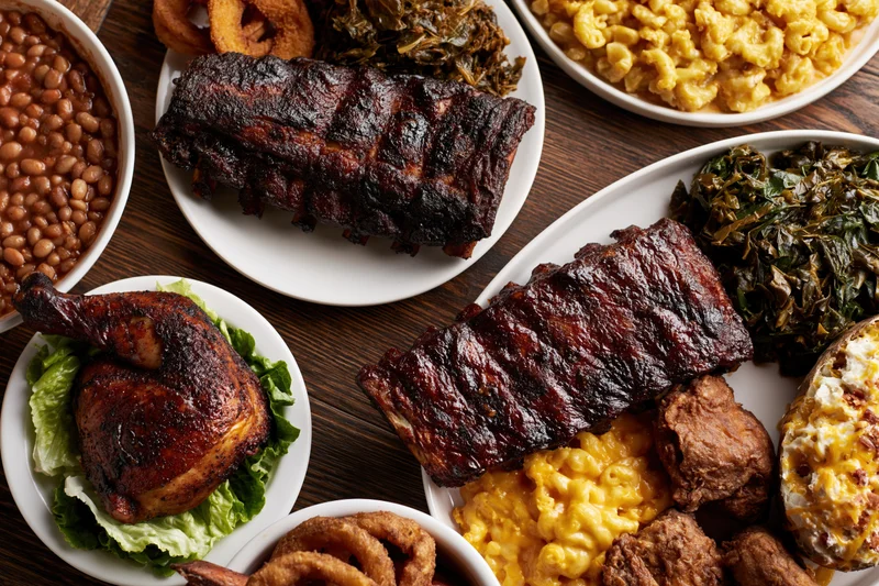 Overhead flat-lay of a southern barbecue spread with ribs, macaroni and cheese, collard greens, baked beans, and smoked chicken on a dark wooden table