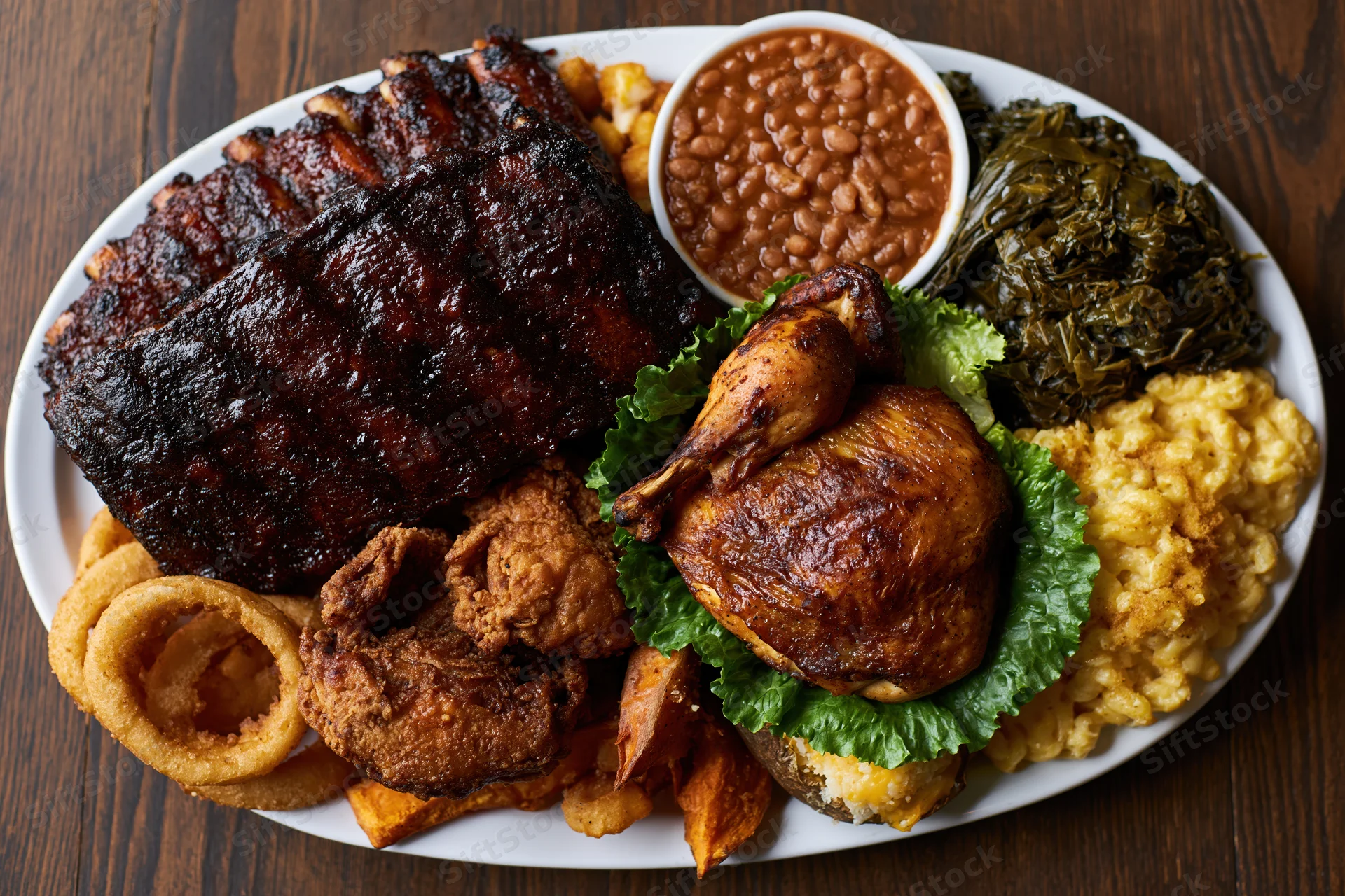 Overhead view of a southern barbecue platter with ribs, chicken, beans, collard greens, macaroni and cheese, onion rings, and sweet potato on a dark wooden table