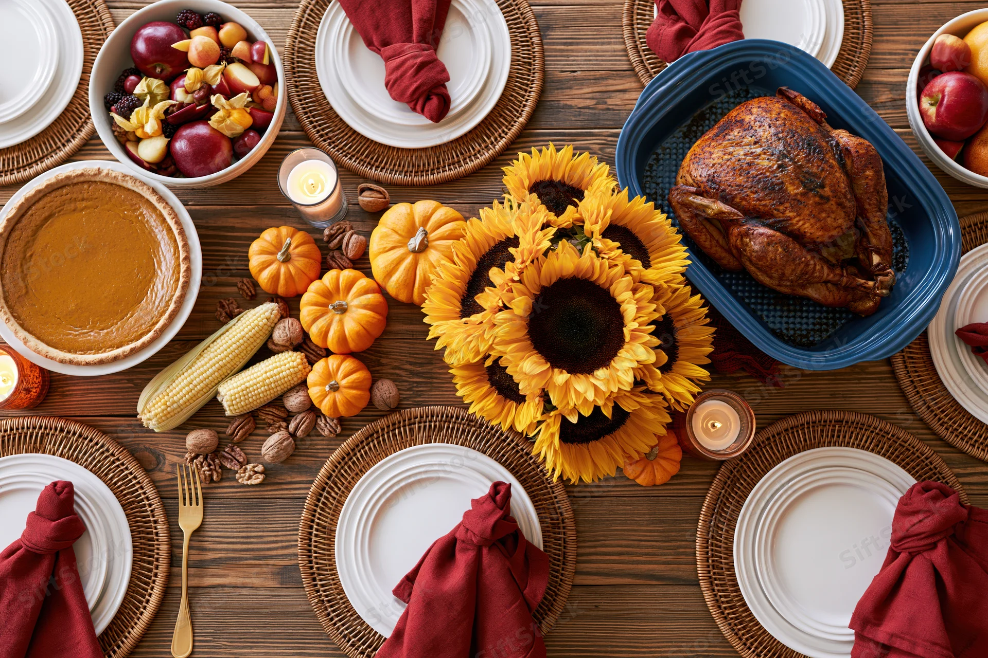 Overhead flat-lay of a thanksgiving dinner table set with turkey, pumpkin pie, sunflowers, corn, and place settings on a wooden surface