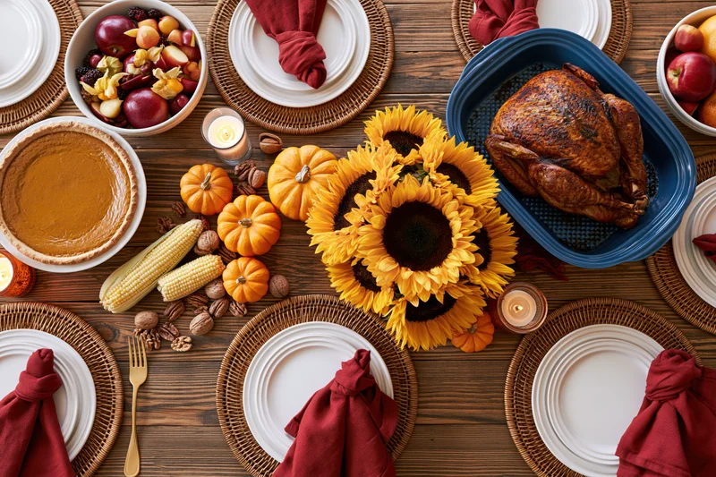 Overhead flat-lay of a thanksgiving dinner table set with turkey, pumpkin pie, sunflowers, corn, and place settings on a wooden surface
