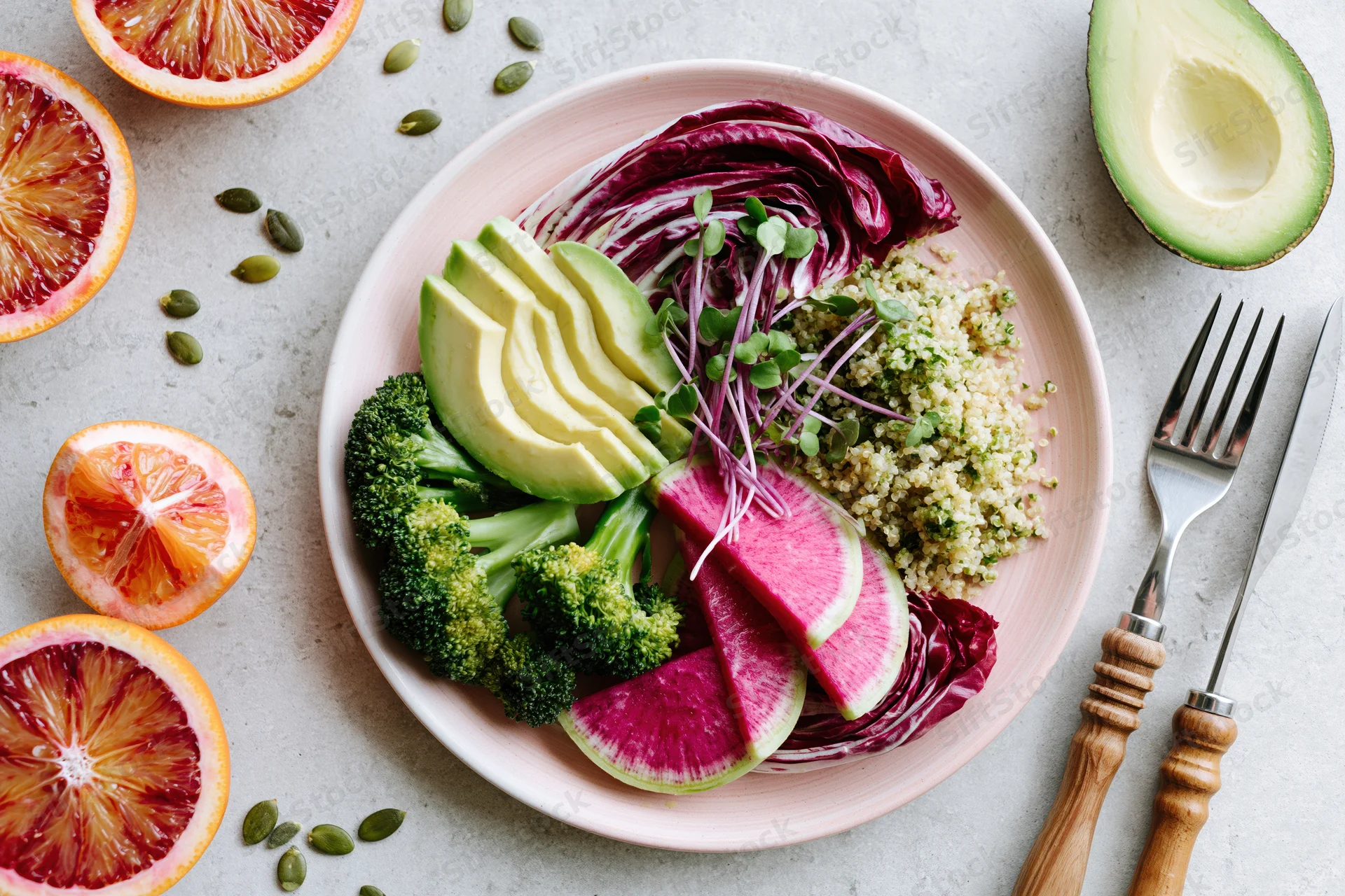 Colorful vegan buddha bowl with avocado, broccoli, quinoa, and blood oranges arranged on a light gray surface with cutlery beside it