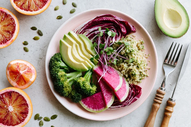 Colorful vegan buddha bowl with avocado, broccoli, quinoa, and blood oranges arranged on a light gray surface with cutlery beside it