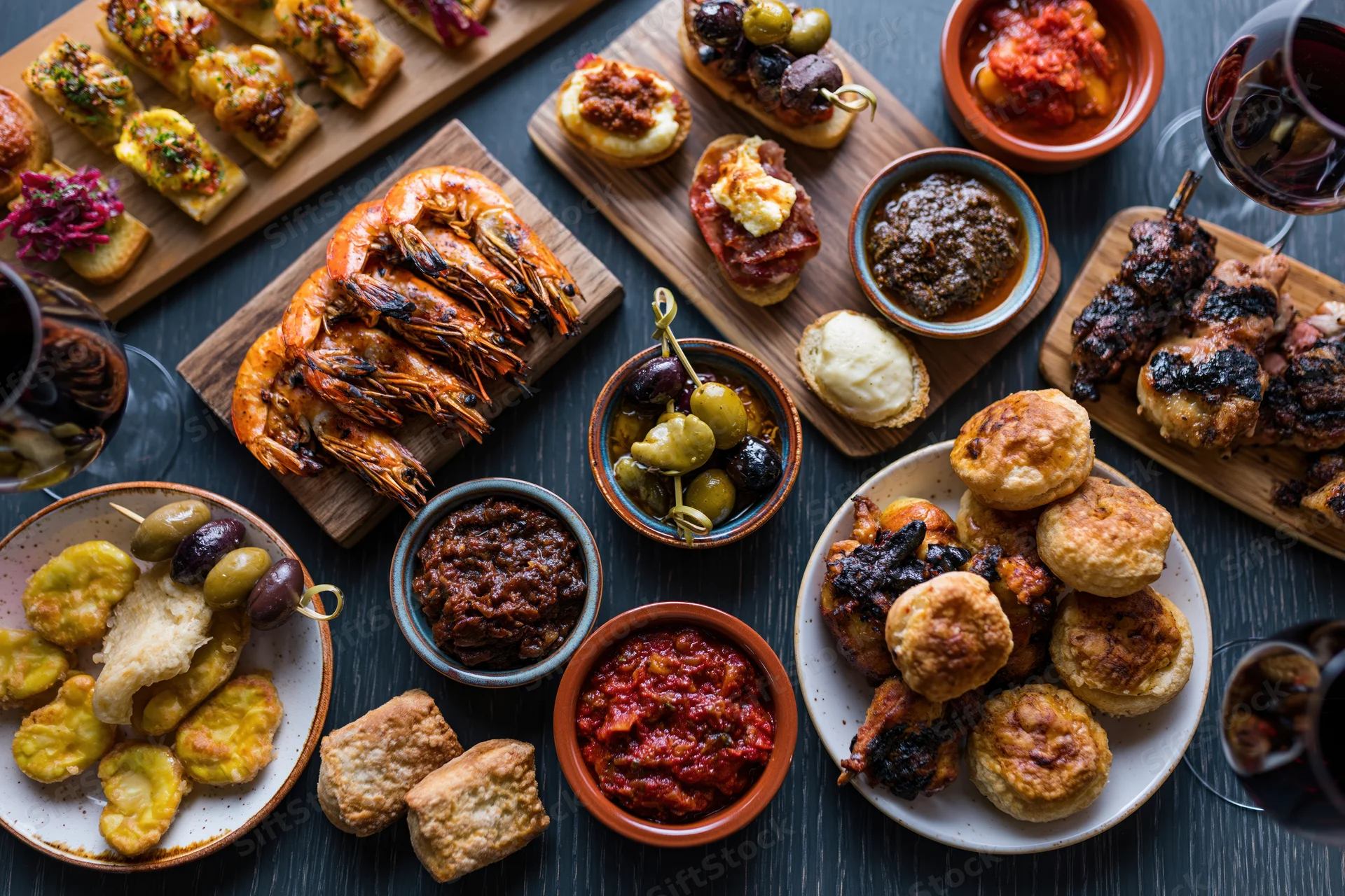 Overhead view of a spanish tapas spread with grilled shrimp, olives, cheese, bread, and wine on a dark wooden table