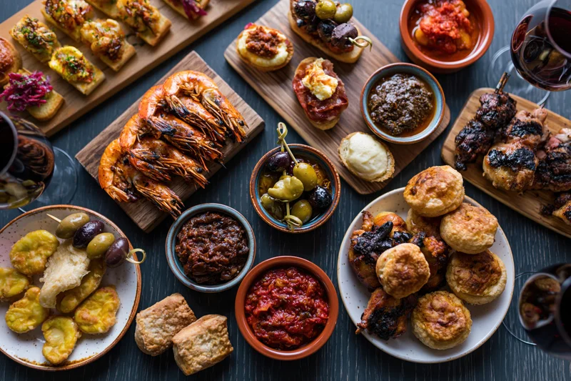Overhead view of a spanish tapas spread with grilled shrimp, olives, cheese, bread, and wine on a dark wooden table