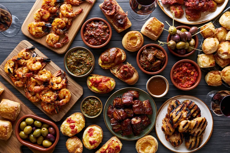 Overhead view of a spanish tapas spread with grilled shrimp, pork, olives, bread, and sauces arranged on a dark wooden table