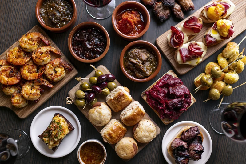 Overhead view of a spanish tapas spread with grilled shrimp, olives, bread, cheeses, sauces and wine on a dark wooden table