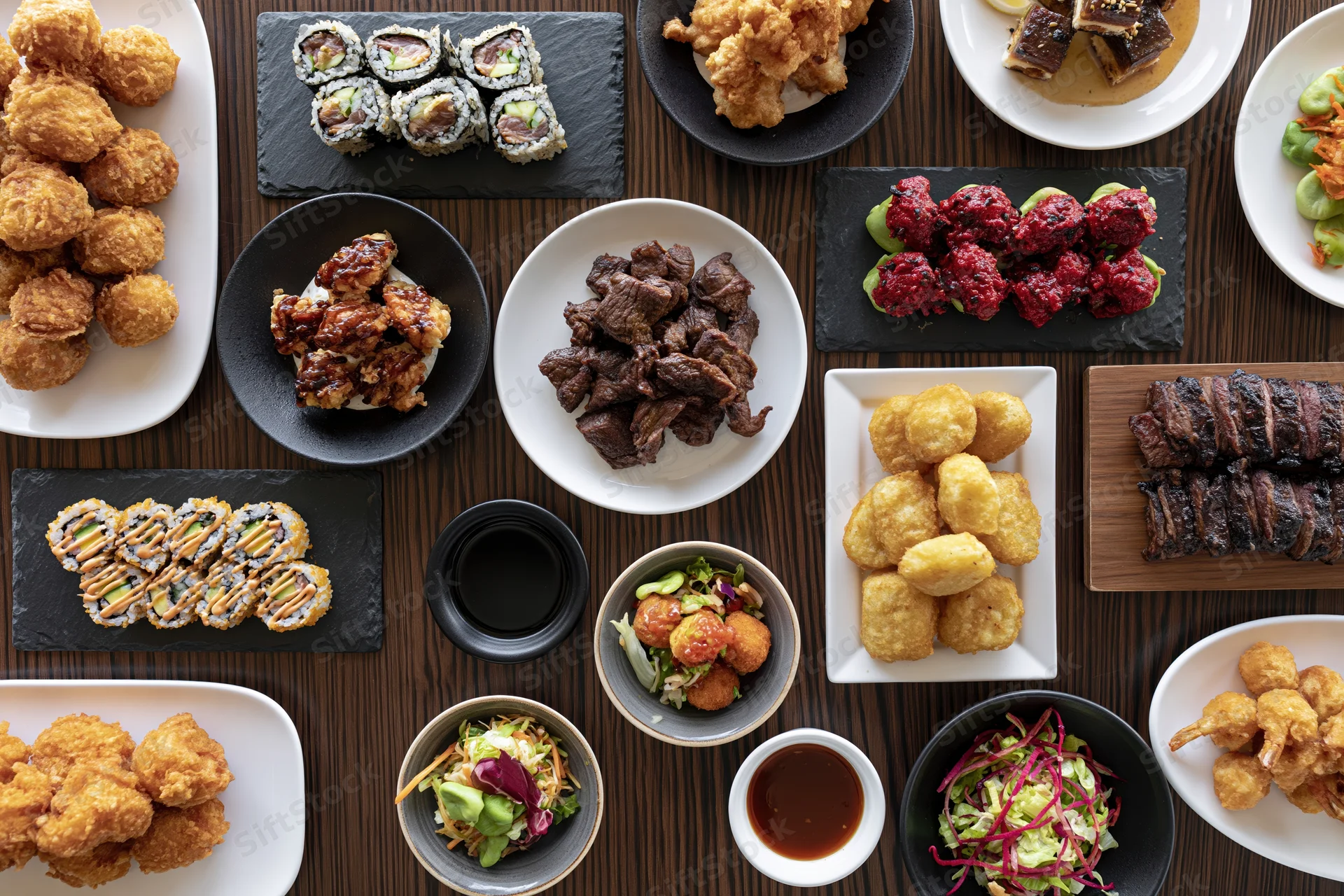 Overhead view of a crowded restaurant table with various Japanese dishes including sushi, tempura, grilled meat, and salads arranged on plates and bowls