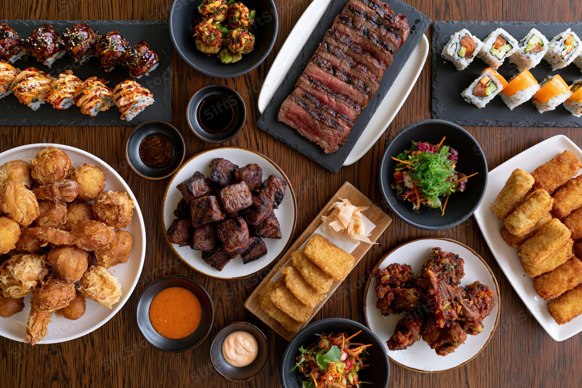 Overhead flat-lay of a restaurant table filled with various dishes including sushi, steak, fried food, and sauces arranged on plates and bowls