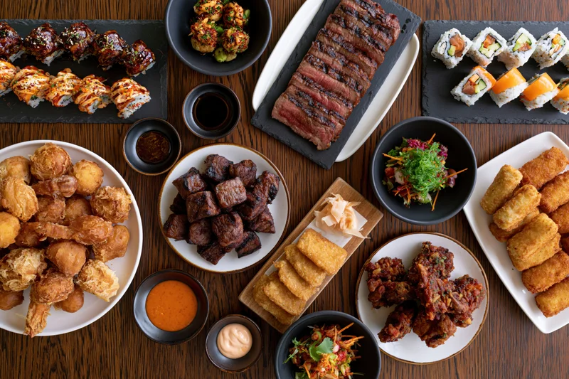 Overhead flat-lay of a restaurant table filled with various dishes including sushi, steak, fried food, and sauces arranged on plates and bowls