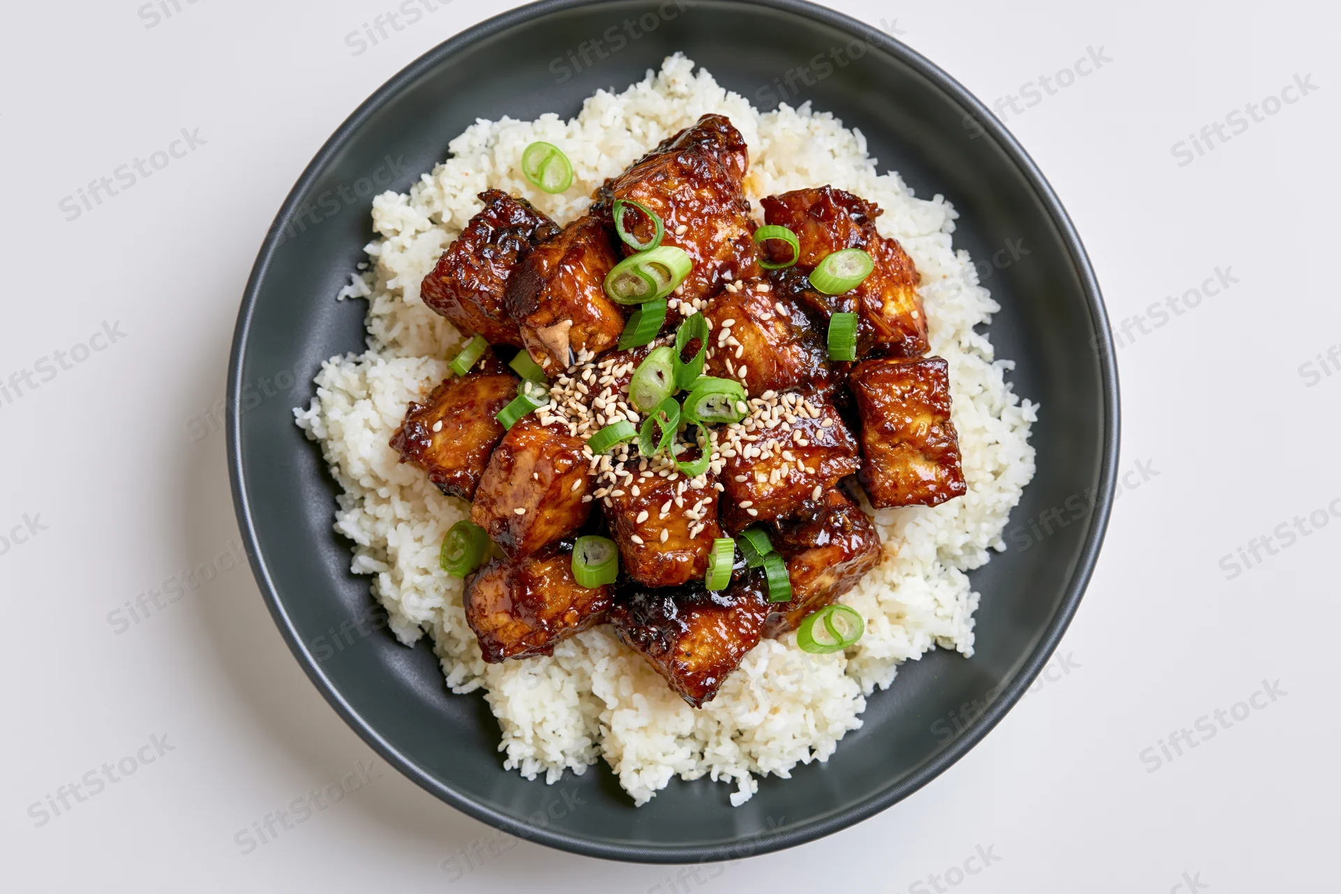 Teriyaki chicken with sesame and scallions served over white rice on a dark matte ceramic plate in overhead flat lay view