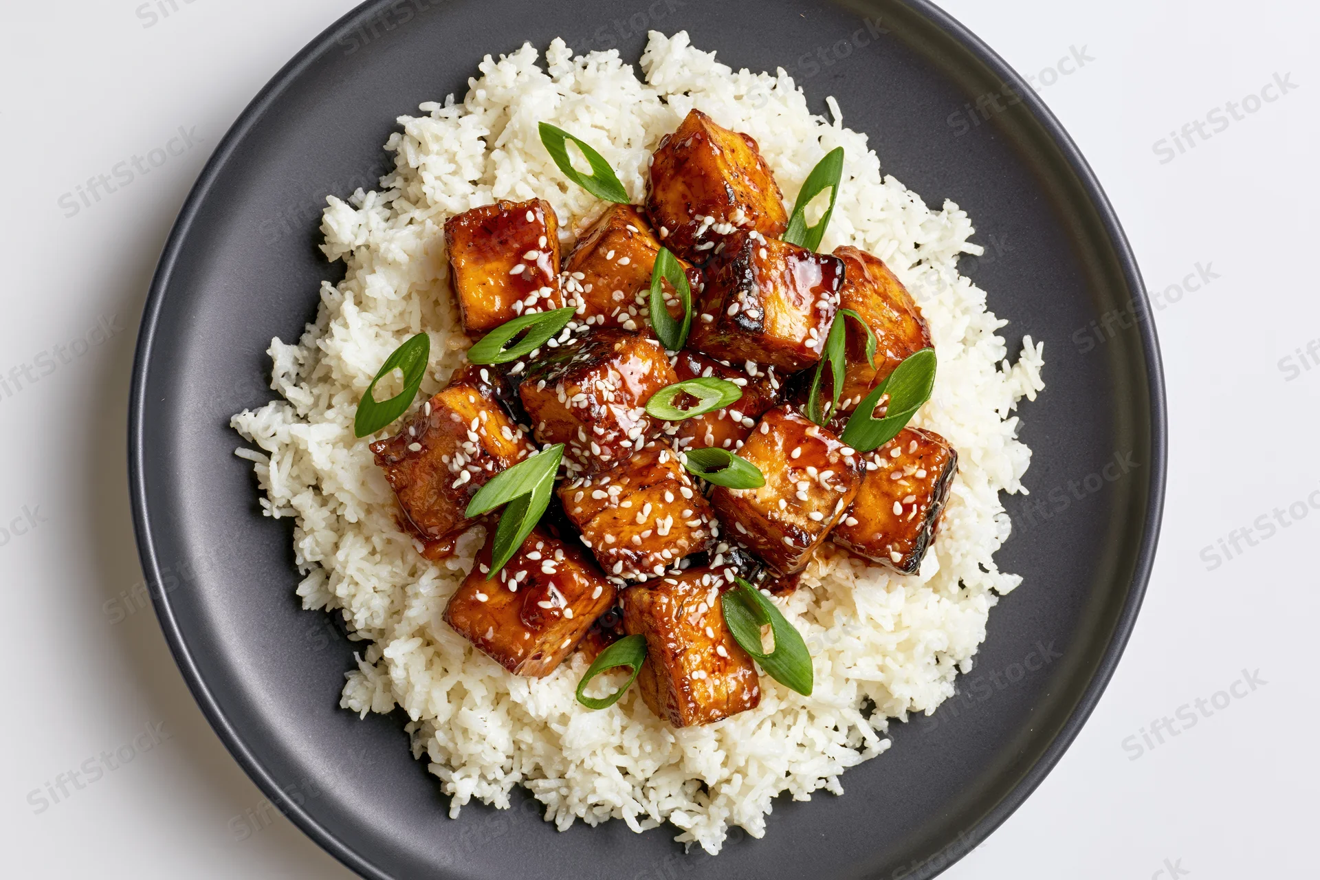 Overhead view of glazed tofu cubes on white rice with sesame seeds and scallions on a dark matte ceramic plate