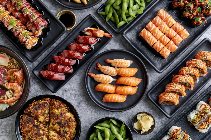 Overhead flat-lay of a large Japanese omakase spread with sushi, sashimi, edamame, and ramen on black plates and trays