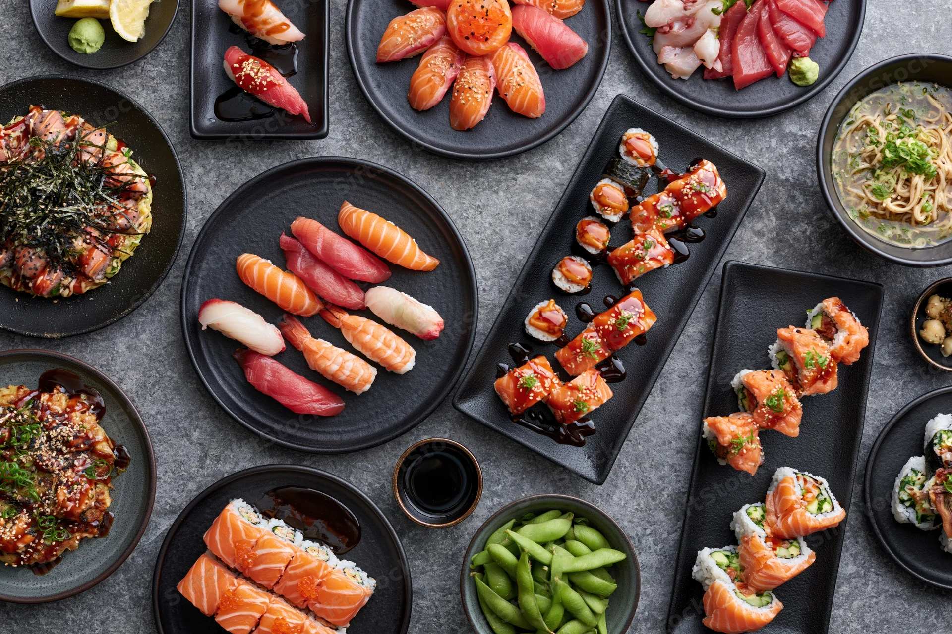 Overhead flat-lay of a large Japanese omakase spread with sushi, sashimi, rolls, and side dishes on black plates arranged on a gray stone table