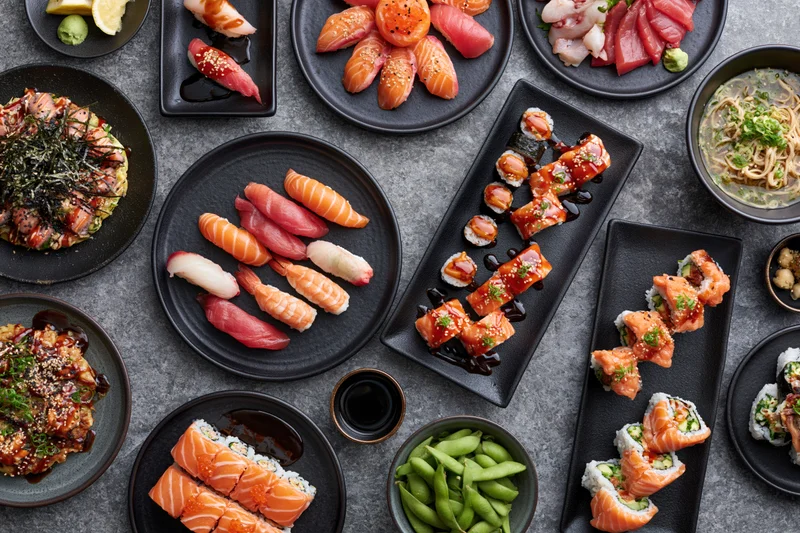 Overhead flat-lay of a large Japanese omakase spread with sushi, sashimi, rolls, and side dishes on black plates arranged on a gray stone table