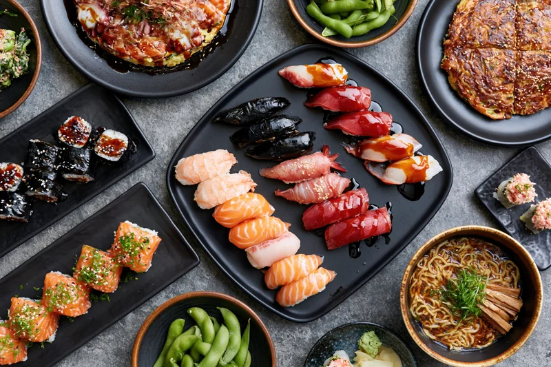 Overhead view of a Japanese omakase spread with sushi, ramen, edamame and garnishes arranged on black plates and bowls on a gray surface