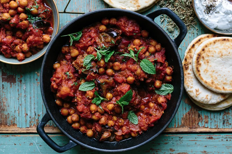 Overhead view of a cast-iron skillet filled with chickpea curry garnished with mint on a rustic blue wooden table with naan and yogurt on the side