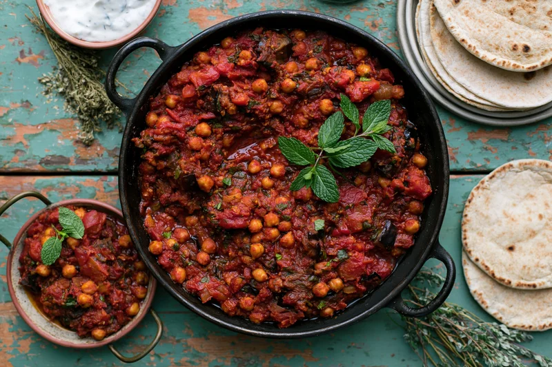Overhead flat-lay of a cast-iron skillet filled with chickpea curry garnished with mint on a distressed teal wooden table with flatbread and yogurt on the side