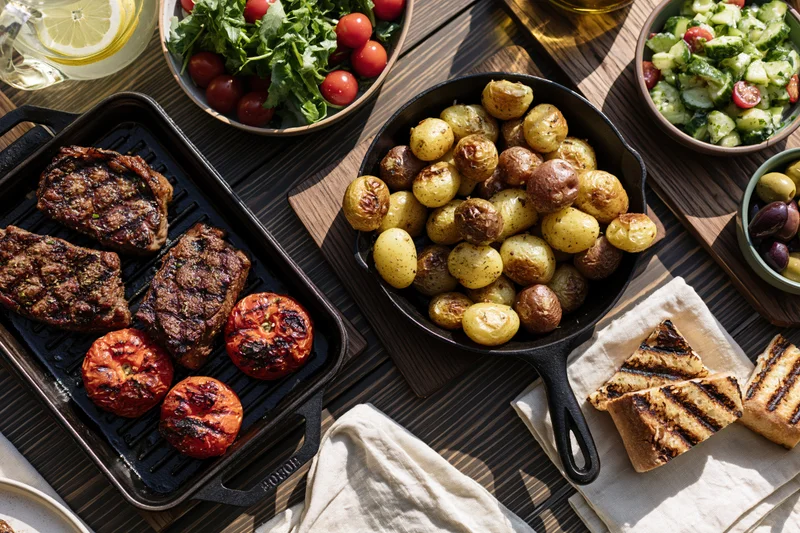 Grilled steak and potatoes with vegetables and bread arranged on a dark wooden table for an outdoor dinner