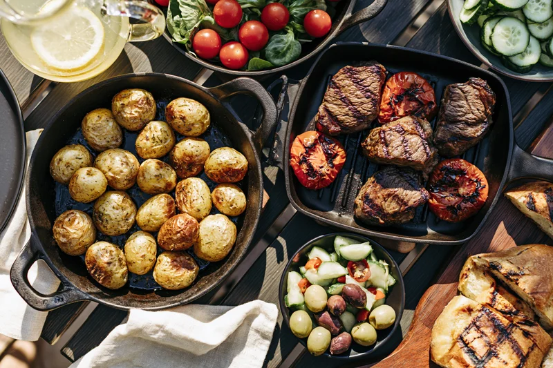 Grilled steak and potatoes with vegetables arranged on a dark wooden table for an outdoor dinner