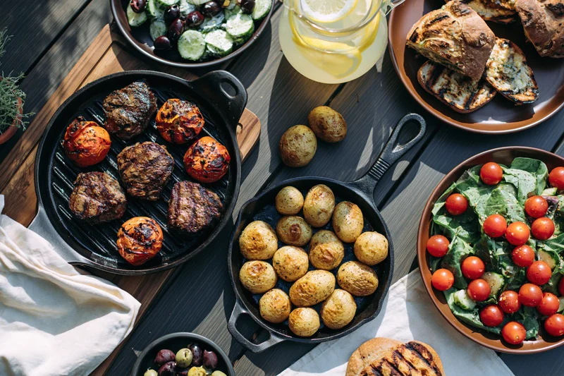 Grilled meat and vegetables in cast iron skillets with salad and bread on a dark wooden table outdoors