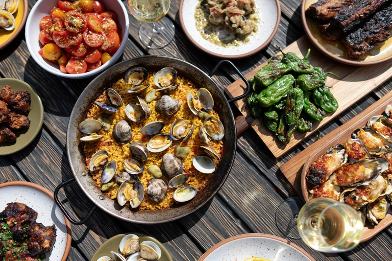 Overhead view of a shared spanish tapas spread with paella, grilled vegetables, seafood, and wine on a wooden table