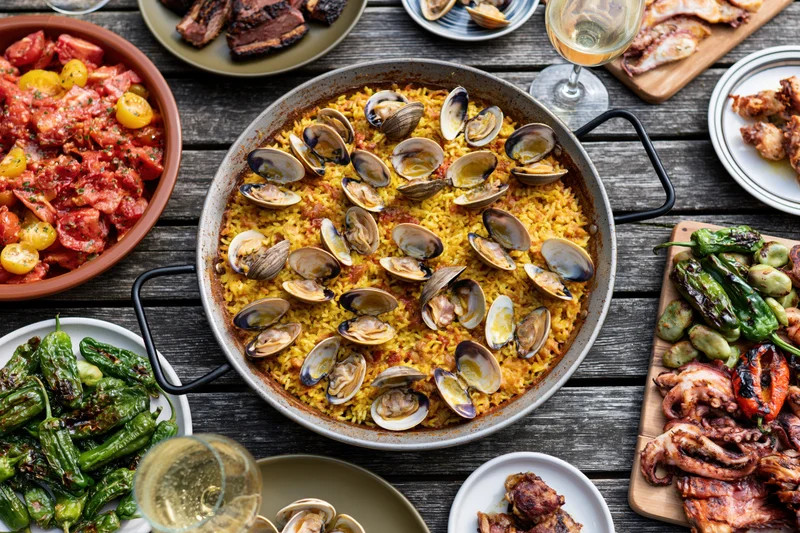 Paella with clams and saffron rice served on a wooden table surrounded by tapas and wine