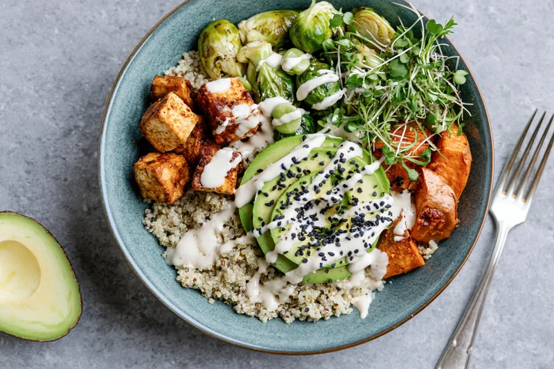 Overhead view of a teal ceramic bowl filled with quinoa, grilled tofu, avocado, brussels sprouts, sweet potato, microgreens, and sesame seeds, drizzled with sauce, on a gray surface with a fork beside it