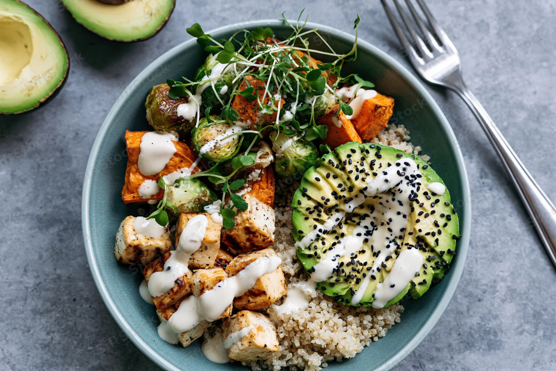 Overhead view of a teal bowl filled with quinoa, grilled tofu, roasted vegetables, avocado, and microgreens, drizzled with sauce and sesame seeds on a gray surface