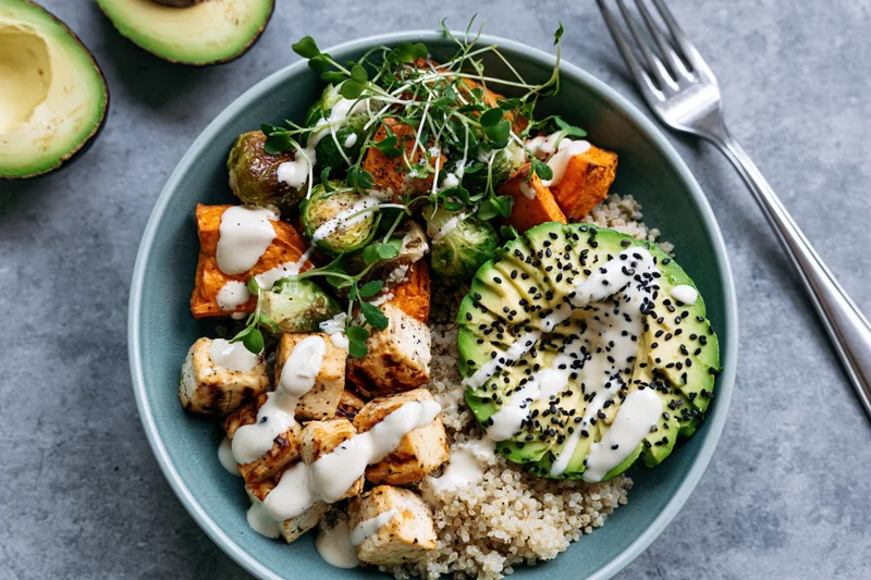 Overhead view of a teal bowl filled with quinoa, grilled tofu, roasted vegetables, avocado, and microgreens, drizzled with sauce and sesame seeds on a gray surface