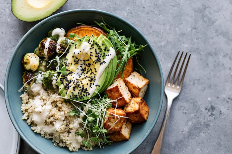 Overhead view of a teal bowl filled with quinoa, avocado, tofu, brussels sprouts and microgreens on a gray surface with a fork beside it