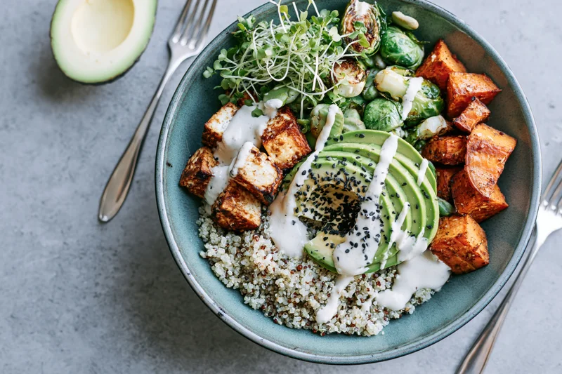 Overhead view of a teal ceramic bowl filled with quinoa, sliced avocado, tofu, roasted sweet potato, brussels sprouts, microgreens and white sauce drizzle on a gray textured surface with forks and half avocado beside it