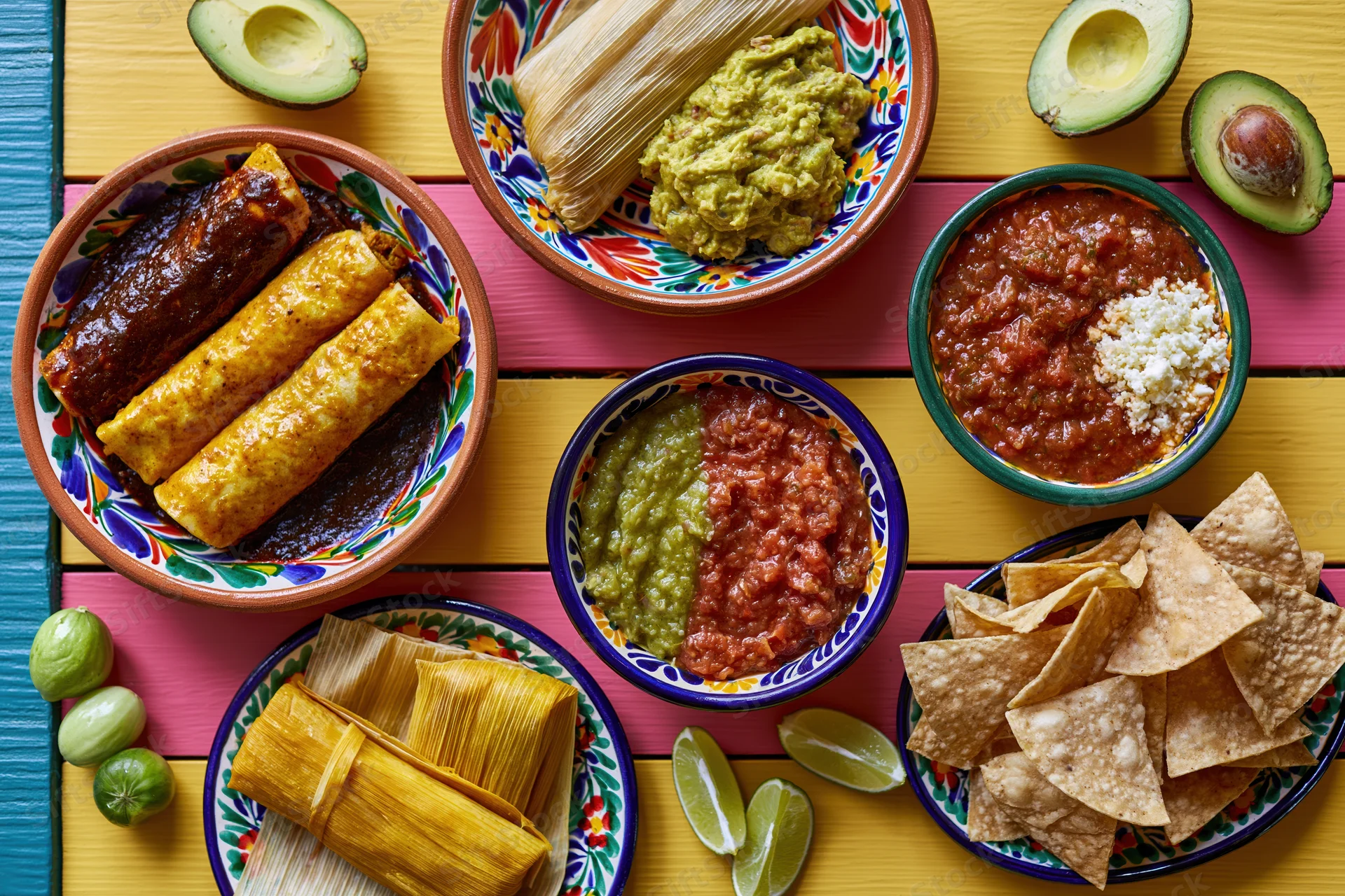 Overhead flat-lay of a traditional mexican food spread with enchiladas, tamales, guacamole, salsa, chips, and avocado on a colorful wooden table